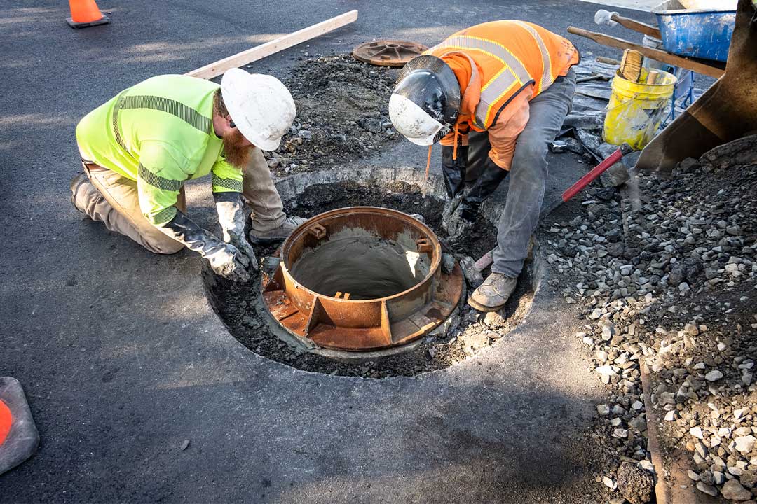 Two workers in hard hats and gloves collaborating on a sewer pipe installation, surrounded by construction materials.